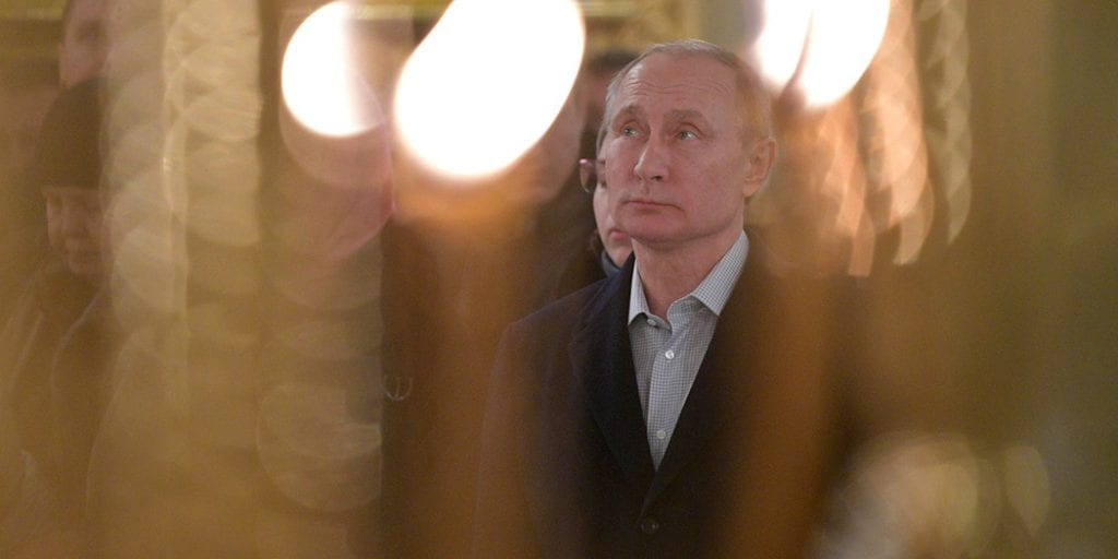 Vladimir Putin looks up as he stands in the middle of a crowd. His surroundings are blurred by candlelight. 