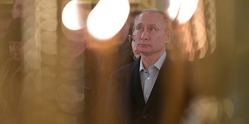 Vladimir Putin looks up as he stands in the middle of a crowd. His surroundings are blurred by candlelight.