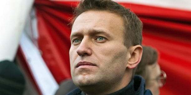 A man with short hair in a dark coat stares into the distance. His background is the flag of Russia.